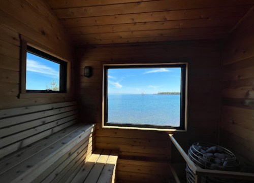 A wooden sauna interior with a view of a calm lake and blue sky through a large window.