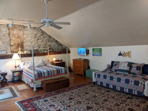 Cozy attic bedroom with a four-poster bed, a daybed, a TV, and rustic stone walls, decorated with a colorful rug.