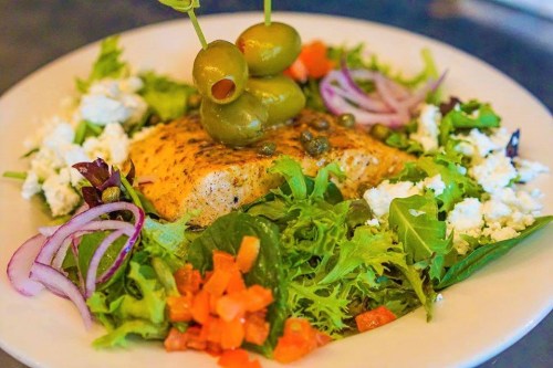 A vibrant salad with grilled salmon, olives, feta cheese, and fresh vegetables on a white plate.
