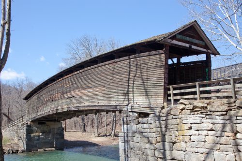 A wooden covered bridge arches over a river, surrounded by bare trees and a clear blue sky.