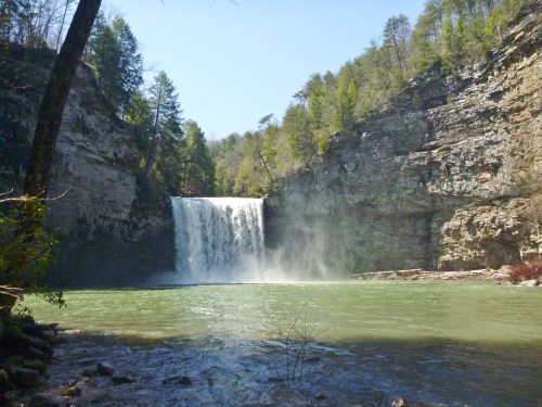 A serene waterfall cascades into a green pool, surrounded by rocky cliffs and lush trees under a clear blue sky.