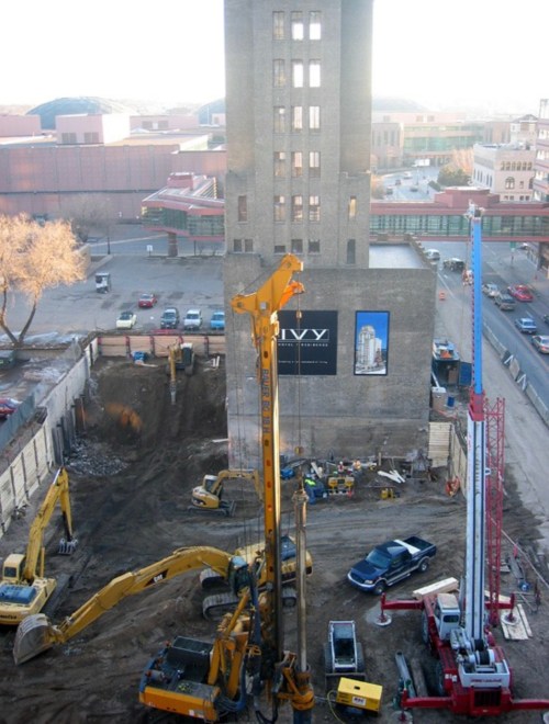 Construction site with heavy machinery, including drills and excavators, near a tall building and parked cars.