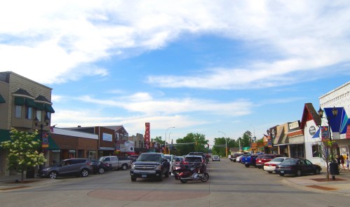A small town street lined with shops and parked cars under a blue sky with scattered clouds.