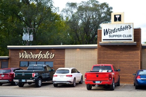 Exterior of Wiederholt's Supper Club with parked cars and a sign featuring a cocktail glass. Trees in the background.