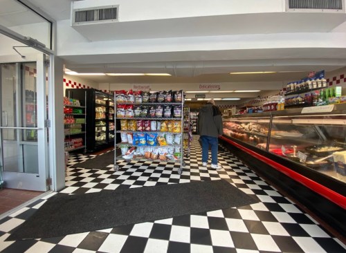 A grocery store interior with checkered floors, shelves of snacks, and a deli counter in the background.