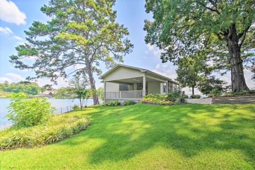 A charming lakeside cottage surrounded by lush greenery and tall trees under a clear blue sky.
