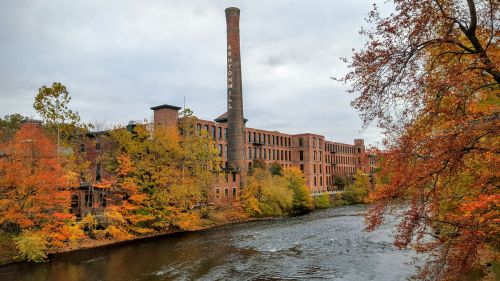 Historic brick factory by a river, surrounded by vibrant autumn foliage and a cloudy sky.