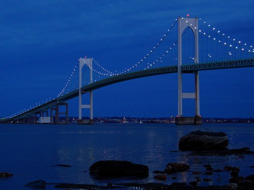 A bridge illuminated at night, spanning over calm waters with a dark blue sky and distant city lights.