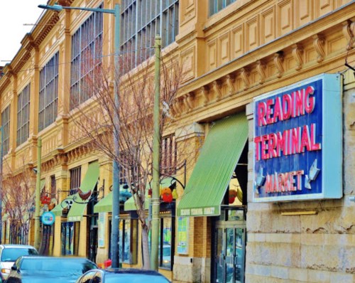 Exterior of Reading Terminal Market with colorful signage and storefronts, featuring trees and parked cars in front.