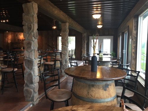 Interior of a rustic restaurant with stone pillars, wooden tables, and empty chairs, featuring large windows.