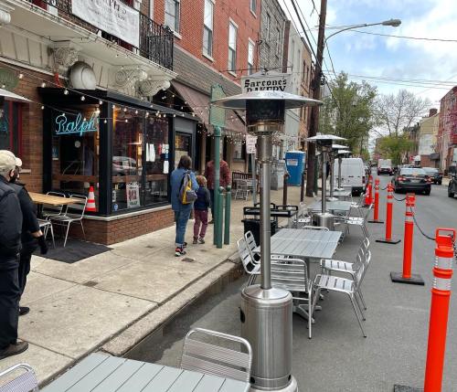 Outdoor dining area with tables and heaters, people walking by, and traffic cones lining the street.