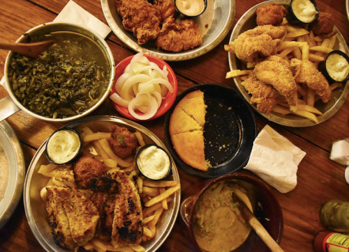 A spread of Southern dishes including fried chicken, cornbread, greens, and fries served in metal trays.