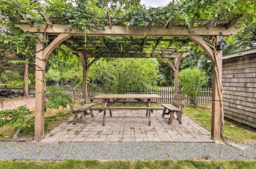 A wooden pergola covered in greenery, with a large picnic table and benches on a stone patio in a garden setting.