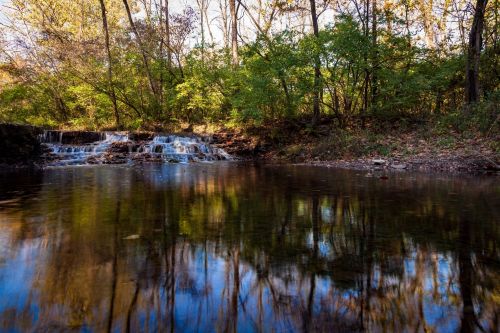 A serene waterfall cascades into a calm pool, surrounded by lush greenery and autumn-colored trees reflecting in the water.