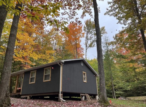 A black cabin surrounded by colorful autumn trees in a serene forest setting.