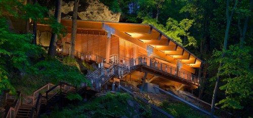 A modern wooden structure illuminated at night, nestled among trees and rocks in a natural setting.
