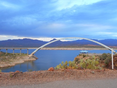 A white arch bridge spans a calm lake, surrounded by rocky shores and mountains under a cloudy sky.