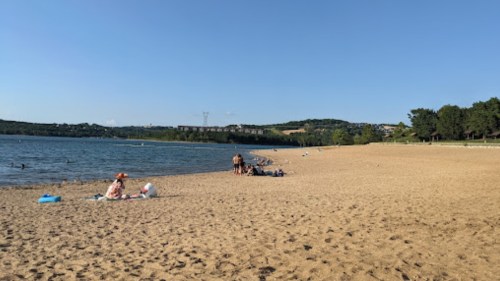 A sandy beach by a lake with people relaxing, swimming, and enjoying the sunny day under a clear blue sky.