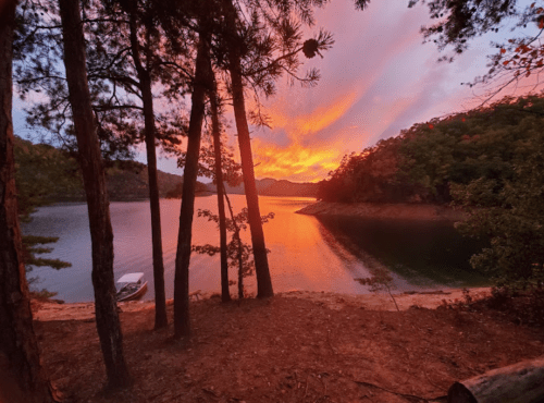 A serene lake at sunset, with vibrant orange and pink skies, framed by tall trees and a boat on the shore.