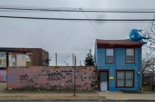 A blue two-story house with graffiti, next to a pink wall, under a cloudy sky. Power lines run overhead.