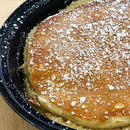 A golden-brown pancake topped with powdered sugar, served in a black dish on a wooden surface.