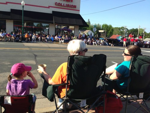 Two women sit in folding chairs on the sidewalk, watching a parade with a crowd gathered across the street.