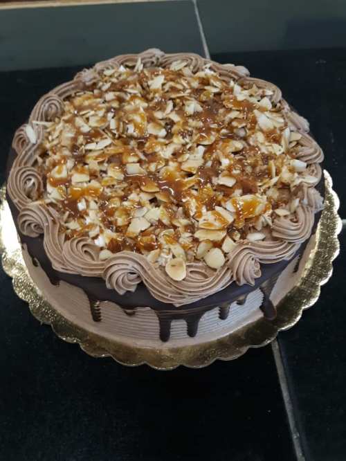 A round chocolate cake topped with caramel, chopped nuts, and decorative frosting on a gold cake board.