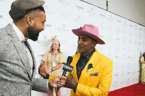 A man in a yellow suit and pink hat speaks to an interviewer on the red carpet at an event, with guests in the background.