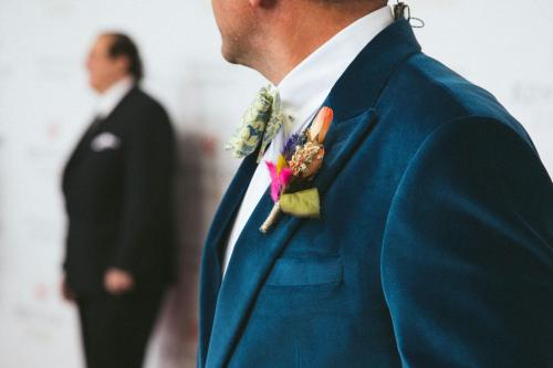 A man in a blue suit with a floral boutonniere stands in focus, while another figure is blurred in the background.
