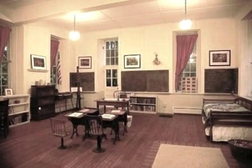 A vintage classroom with wooden desks, chalkboards, bookshelves, and large windows, featuring warm lighting and historical decor.