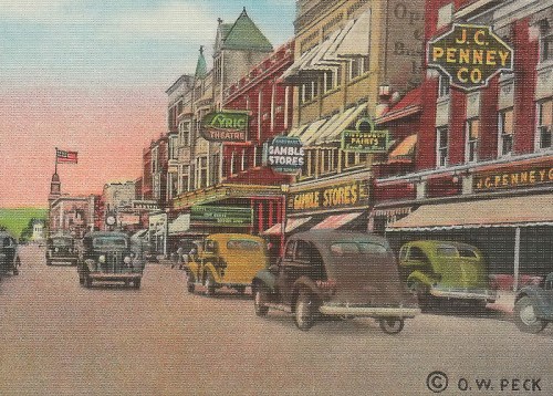 Historic street scene with vintage cars and storefronts, featuring J.C. Penney and a theater, under a pastel sky.