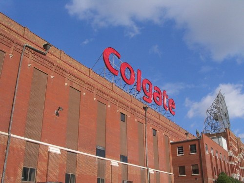 Red "Colgate" sign on a brick building under a blue sky with clouds.
