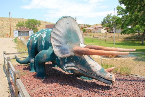 A large, colorful triceratops statue in a grassy area, surrounded by rocks and a playground in the background.