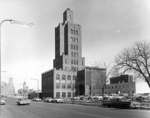 Historic tall brick building with a tower, surrounded by parked cars and trees, in a cityscape. Black and white photo.
