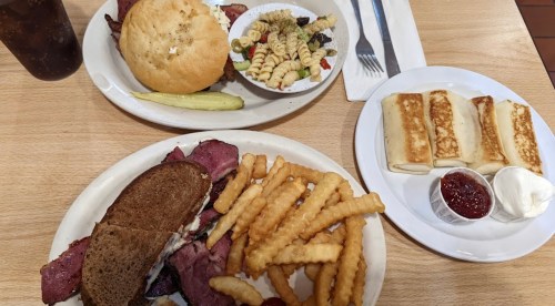 A plate with a sandwich, crinkle-cut fries, and a side of pasta salad, alongside a burger and dessert rolls.