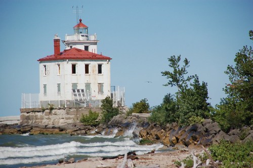 An old lighthouse stands by the shore, surrounded by waves and trees under a clear blue sky.