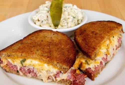 A toasted sandwich filled with corned beef and cheese, served with a side of creamy coleslaw and a pickle.