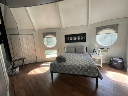 Cozy bedroom with a round window, gray bedding, wooden floors, and modern decor, illuminated by natural light.