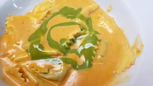 A bowl of ravioli topped with a creamy orange sauce and a drizzle of green sauce.