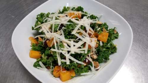 A fresh salad with kale, butternut squash, nuts, and topped with shredded cheese, served in a white bowl.