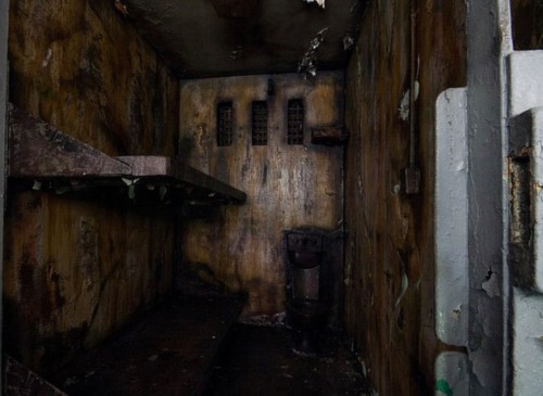 A dark, abandoned cell with peeling walls, rusty shelves, and a toilet in the corner, evoking a sense of decay.