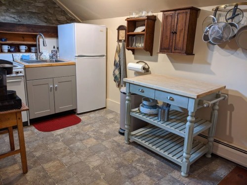 Cozy kitchen with a fridge, sink, wooden table, and shelves, featuring a rustic design and stone wall accents.