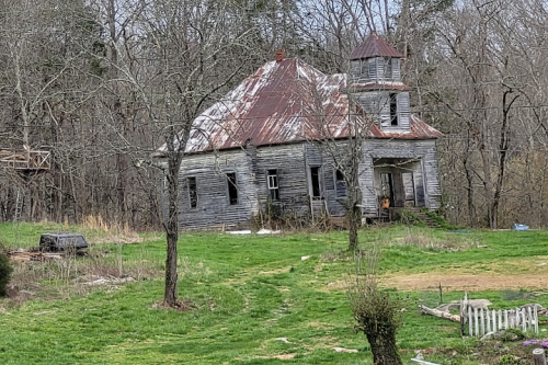Abandoned, weathered house with a rusty roof, surrounded by trees and overgrown grass.