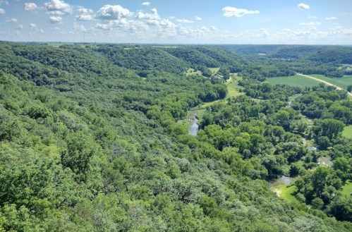A panoramic view of lush green hills and a winding river under a bright blue sky with scattered clouds.