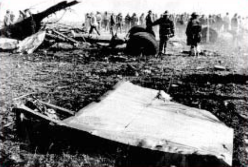 A black and white image of a plane crash site with debris and people surveying the area in the background.