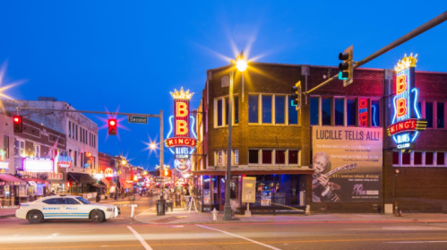 A vibrant street scene at dusk featuring BB King's Blues Club, neon lights, and a police car parked nearby.