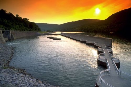 A serene river at sunset, with a dam and circular spillways reflecting the warm colors of the sky.