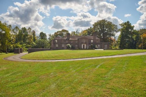A large, elegant brick house surrounded by lush green grass and trees under a partly cloudy sky.