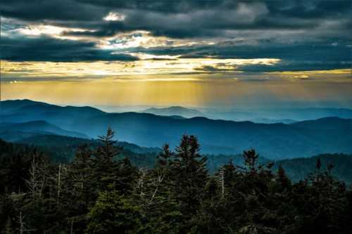 A panoramic view of layered mountains under a dramatic sky, with sunlight breaking through clouds and illuminating the landscape.