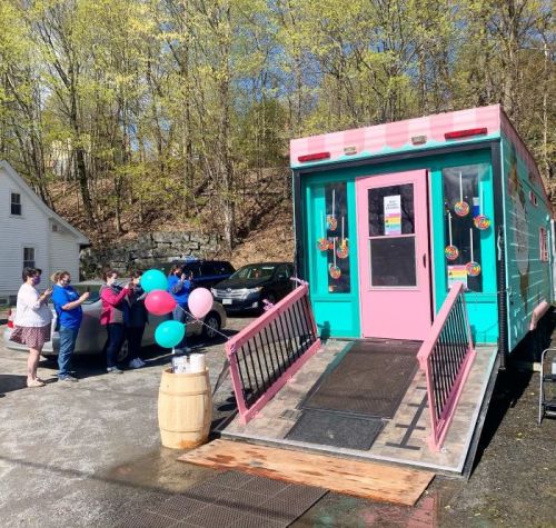 A colorful food truck with balloons outside, surrounded by people enjoying the sunny day.